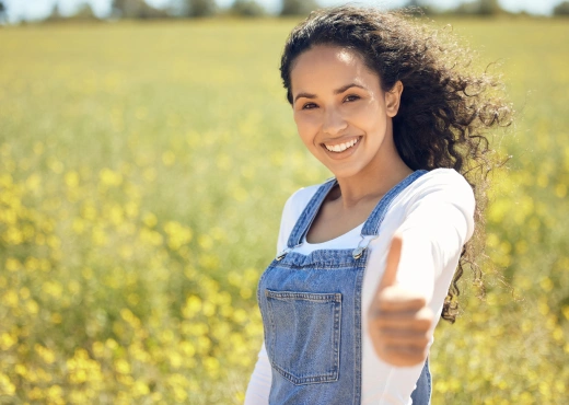 A young woman joyfully giving a thumbs-up in a field of flowers, representing the positive mindset encouraged in the Happiness Program.